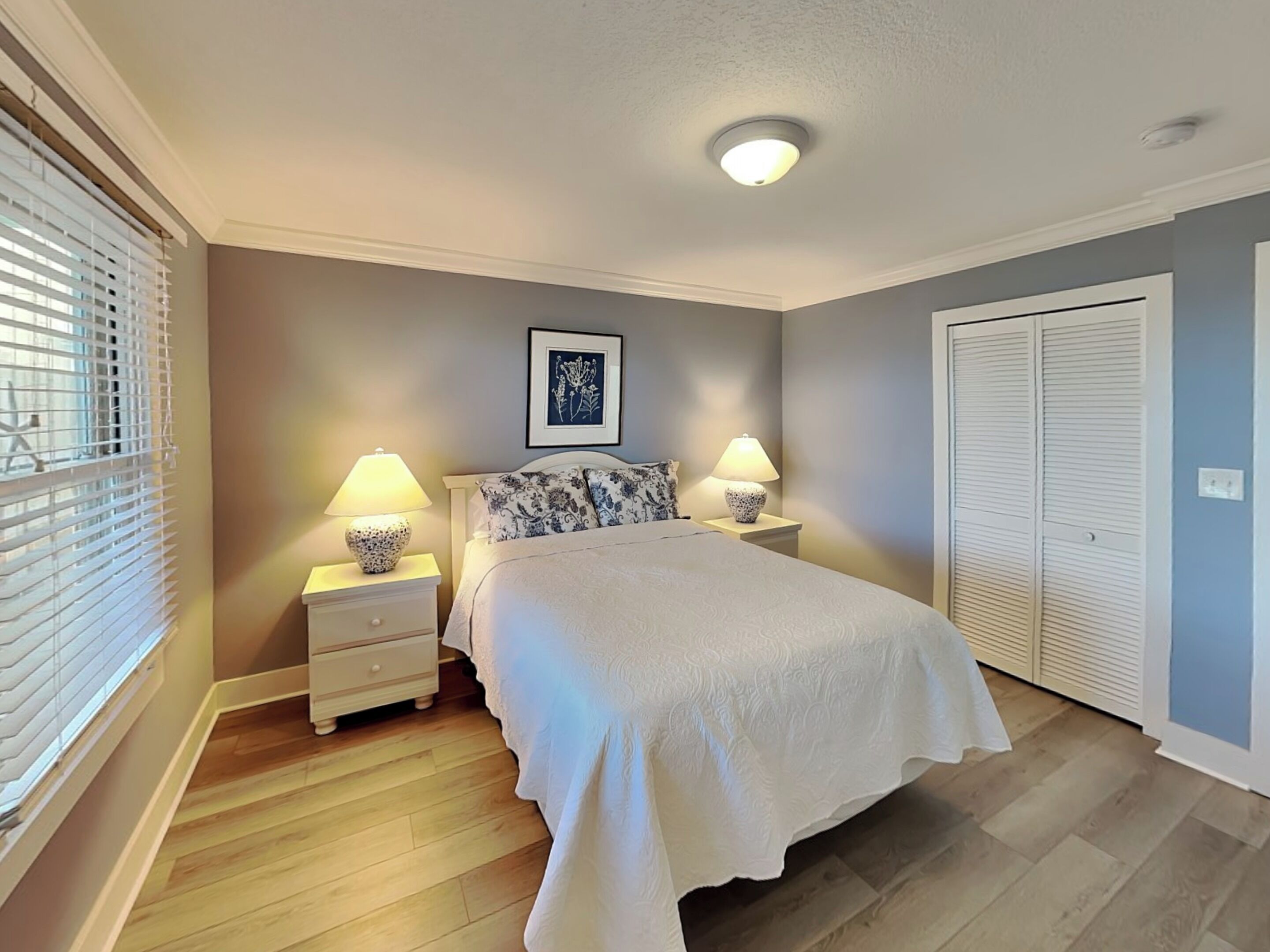 A bedroom with a bed, two bedside tables with lamps, a ceiling light, a window with blinds, and a door.