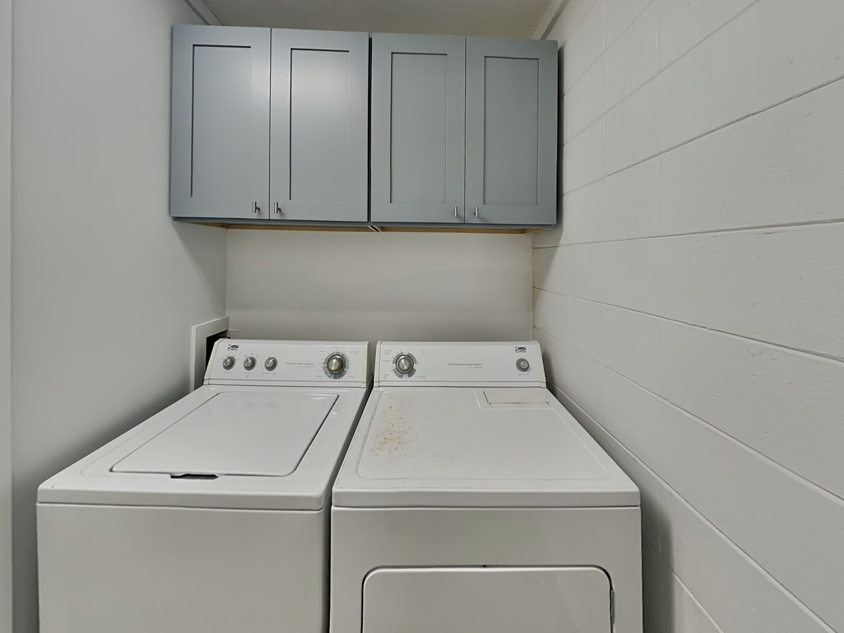 A laundry room with two white washing machines and grey cabinets above.