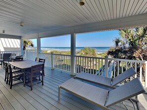 A covered outdoor area with a table and chairs, a lounge chair, and a view of the beach and ocean.