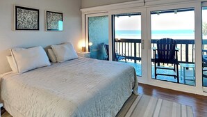 A bedroom with a large bed, a nightstand, two framed artworks, and a view of the beach through a sliding glass door. - Island House by Distinctive Beach Rentals (St. Augustine)