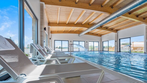 An indoor swimming pool with clear views of the outside, wooden ceiling beams, and lounge chairs.