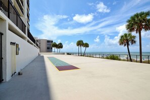 A beachfront walkway with a shuffleboard court, palm trees, and a building with balconies.
