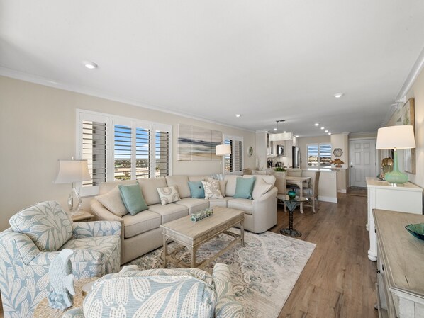 A spacious living area with a sofa set, a coffee table, and a dining area with a kitchen in the background.