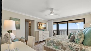Ein Schlafzimmer mit Bett, Schreibtisch, Stuhl, Kommode, Deckenventilator, einem Gemälde und Blick auf den Ozean.