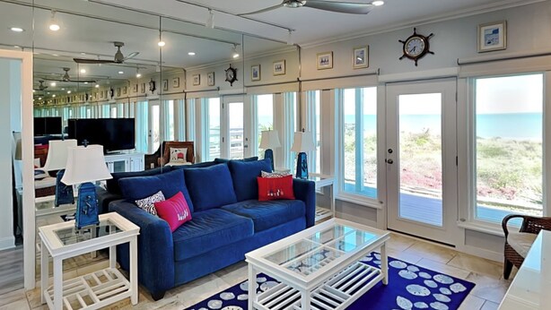 A spacious living room with a blue sofa, white coffee table, and a large window offering a view of the beach. - Ocean Pearl by Distinctive Beach Rentals (St. Augustine)