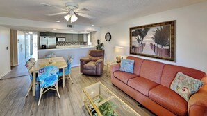 A modern living room with a red sofa, a glass coffee table, and a dining area with a wooden table and chairs. - Ocean Villas 107 by Distinctive Beach Rentals (St. Augustine)