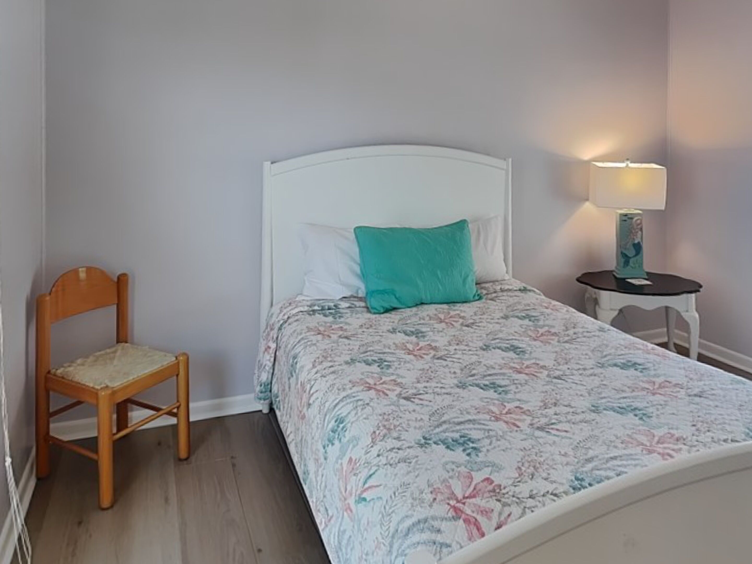 Une chambre à coucher avec un lit, une chaise en bois, une table de chevet avec une lampe et une étagère fixée au mur.