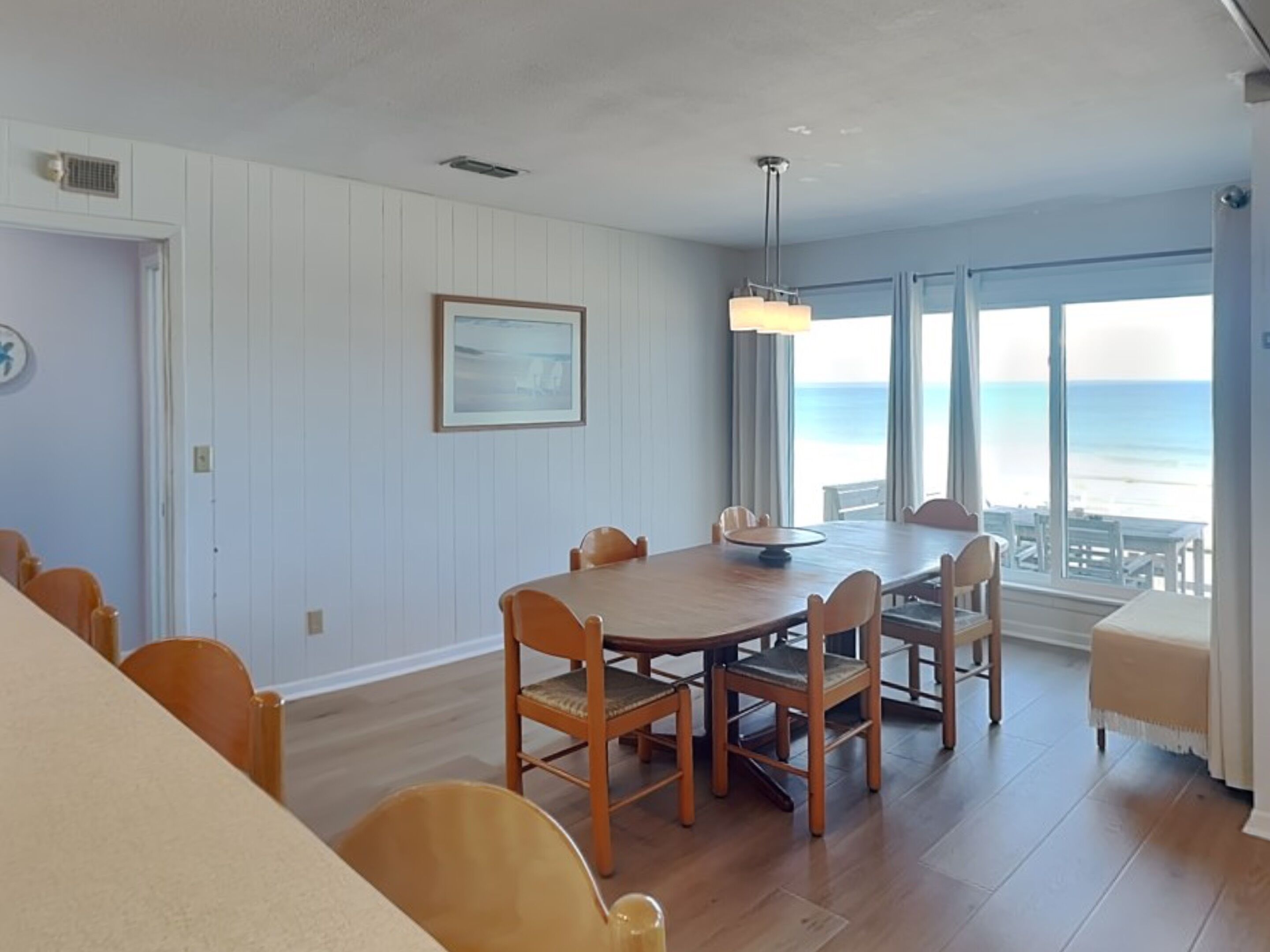 A dining area with a wooden table and chairs, a pendant light, and a view of the ocean through large windows.