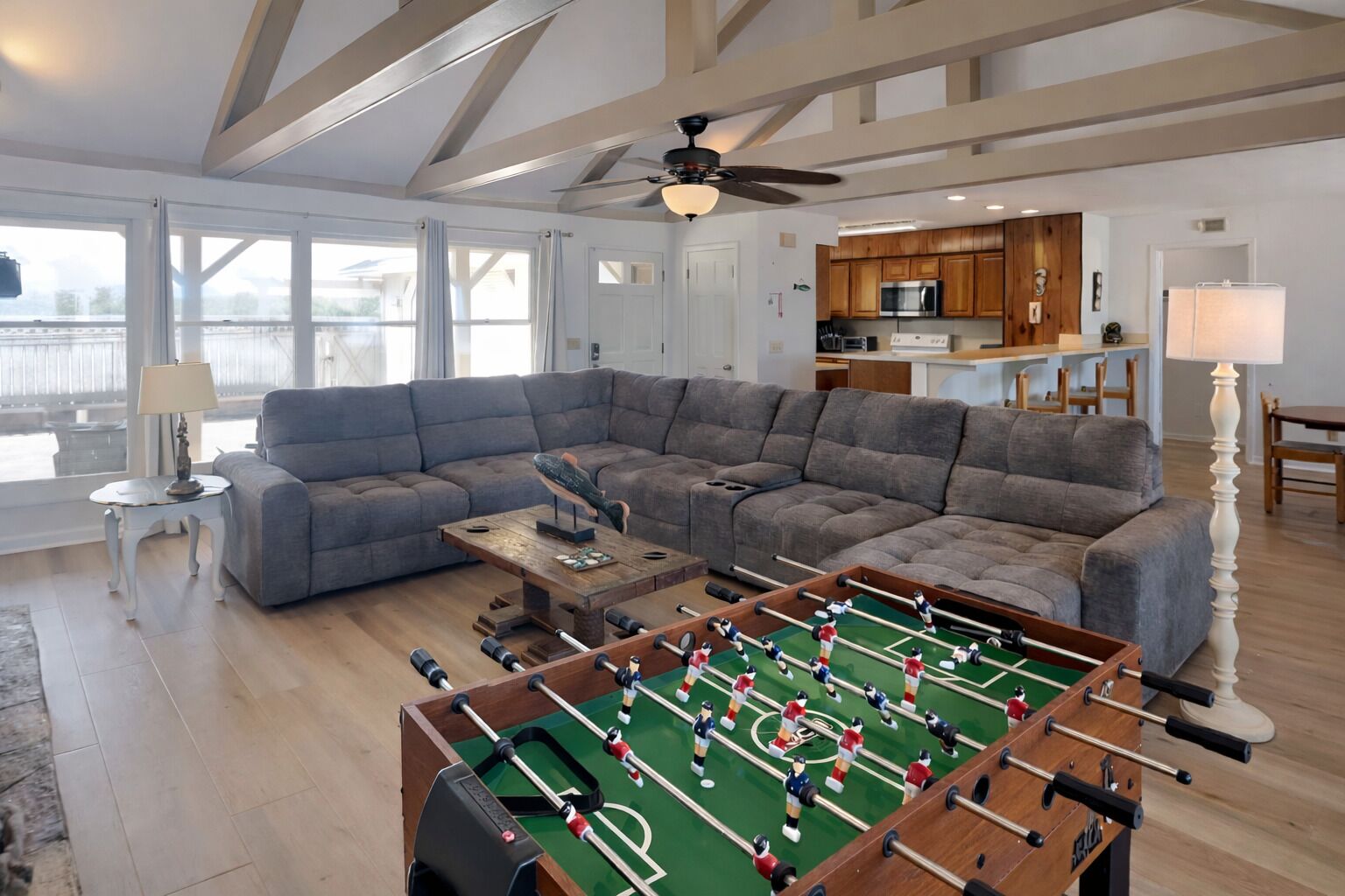 A spacious living room with a large sectional sofa, a foosball table, and a kitchen area with wooden cabinets and a ceiling fan.