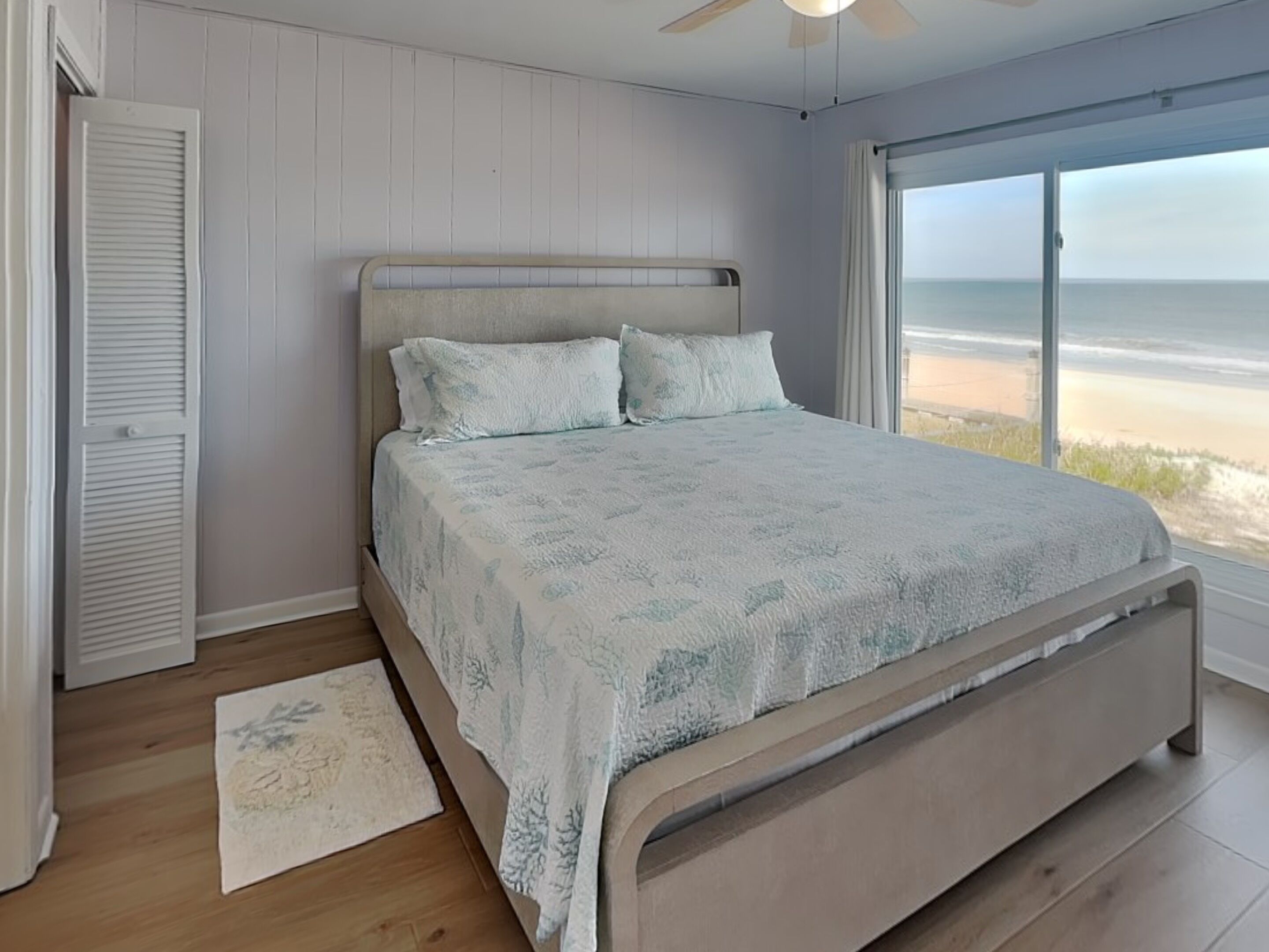 Un dormitorio con cama, ventilador de techo, vista a la playa y suelo de madera.