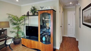 A living room with a wooden entertainment center, a flat-screen TV, a potted plant, and a dining area with a table and chairs. - Sea Urchin 39 by Distinctive Beach Rentals (St. Augustine)