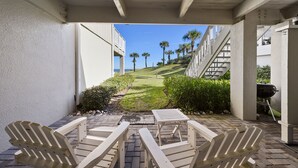 Un patio con sillas y mesa de madera, una parrilla y vistas a una zona ajardinada con palmeras.