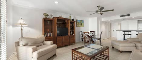 A living room with a beige sofa, a wooden entertainment unit, a glass coffee table, and a ceiling fan.