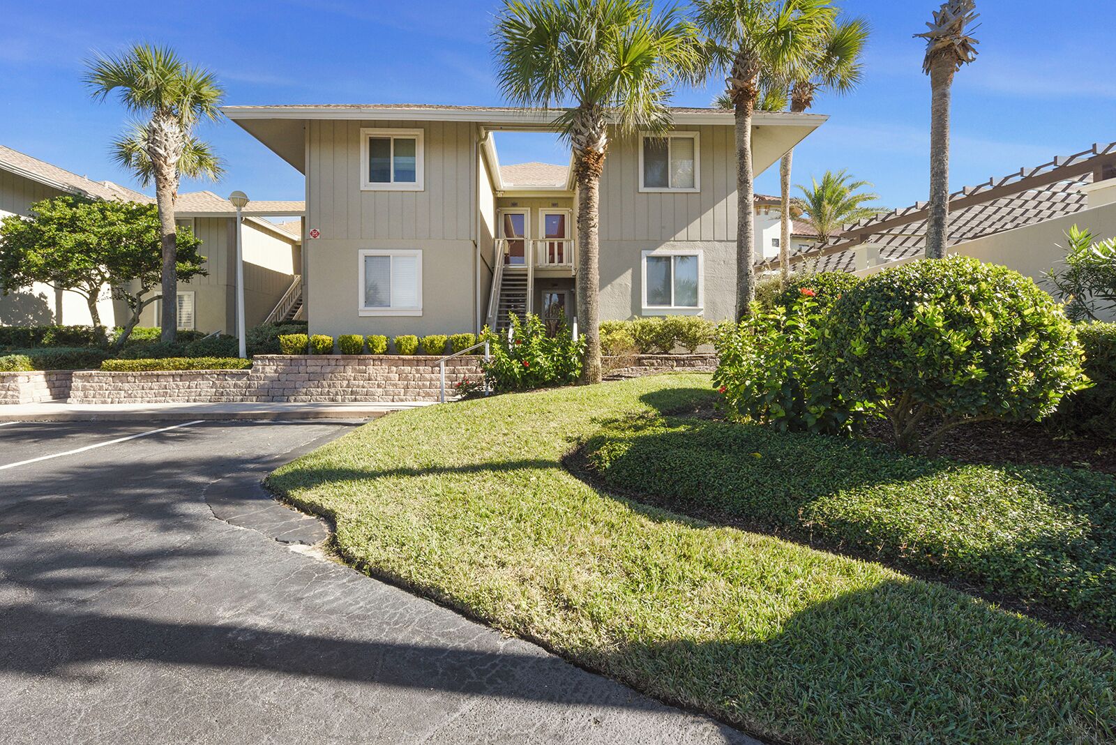 A two-story residential building with a front entrance, a stone retaining wall, and a well-maintained lawn with palm trees.