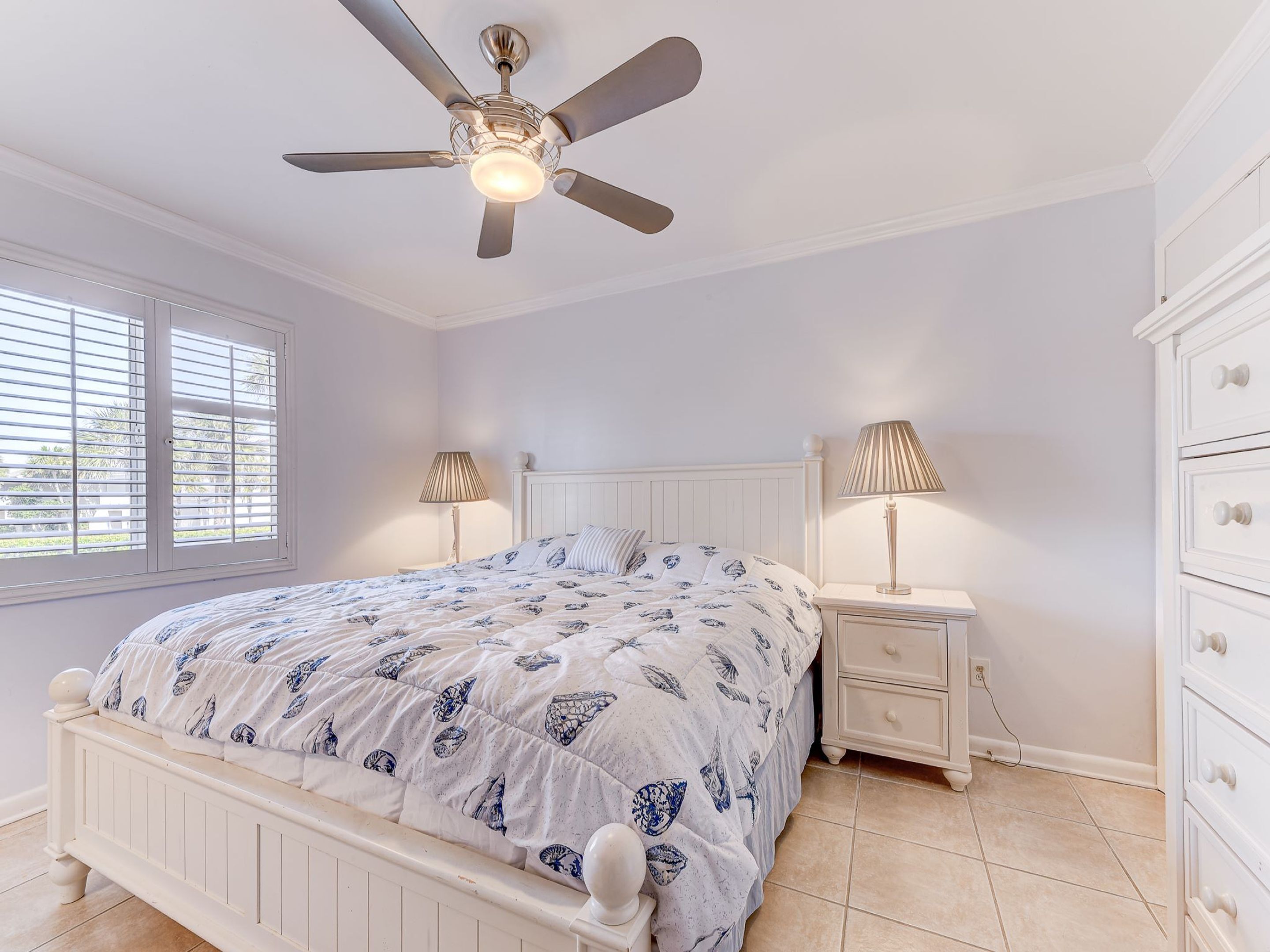 Une chambre à coucher comprenant un lit, des tables de chevet, une commode, un ventilateur de plafond et une fenêtre avec des stores.
