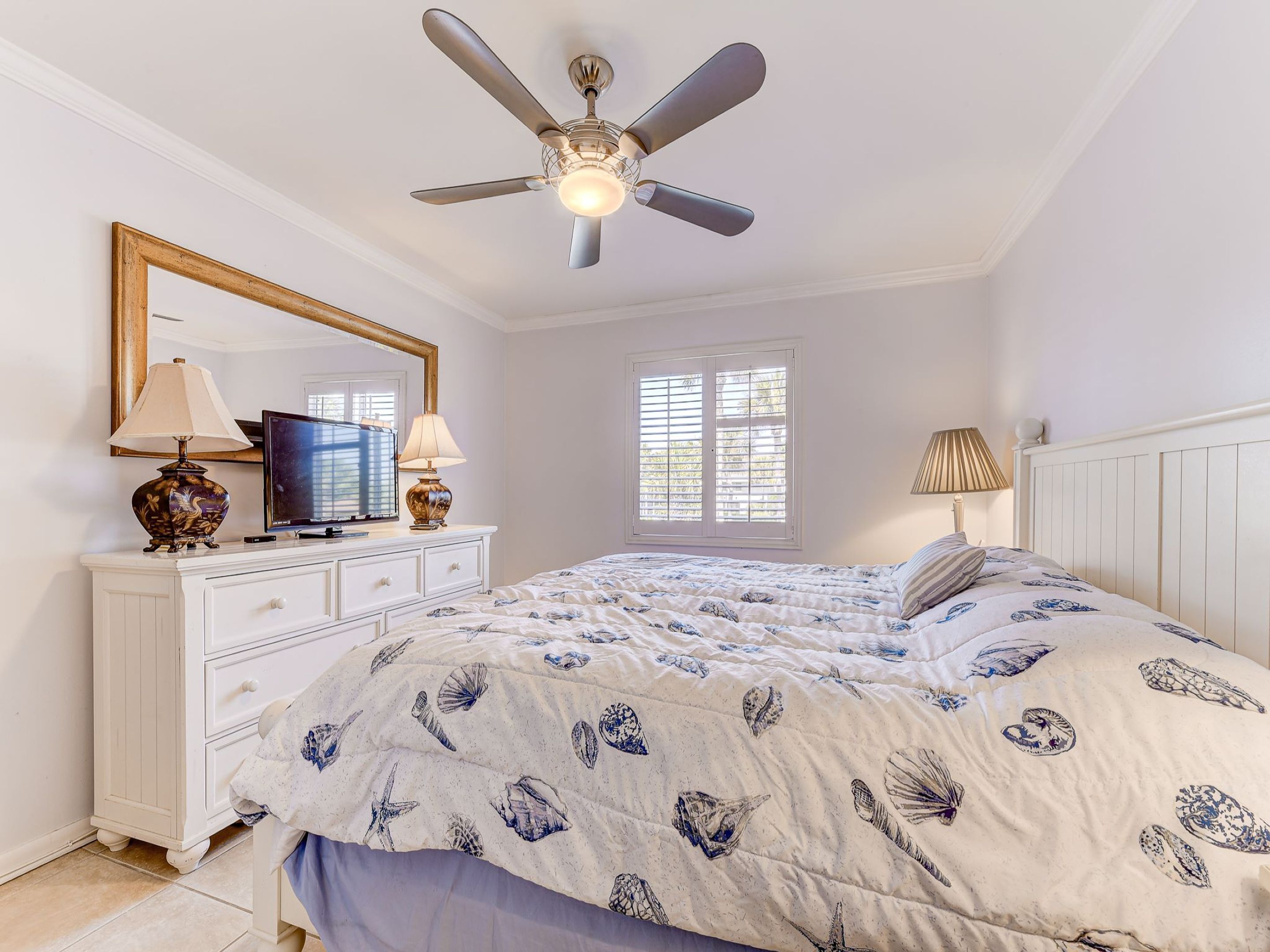 Une chambre à coucher avec un lit, un ventilateur de plafond, un téléviseur posé sur une commode et une fenêtre avec des stores.