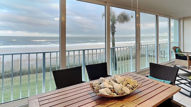 Un balcon donnant sur la plage, une table en bois sur laquelle est posé un bol rempli de coquillages, et un ventilateur de plafond.