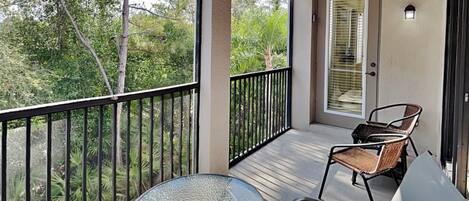 A balcony with a glass table and two chairs, a door with blinds, and a view of trees.