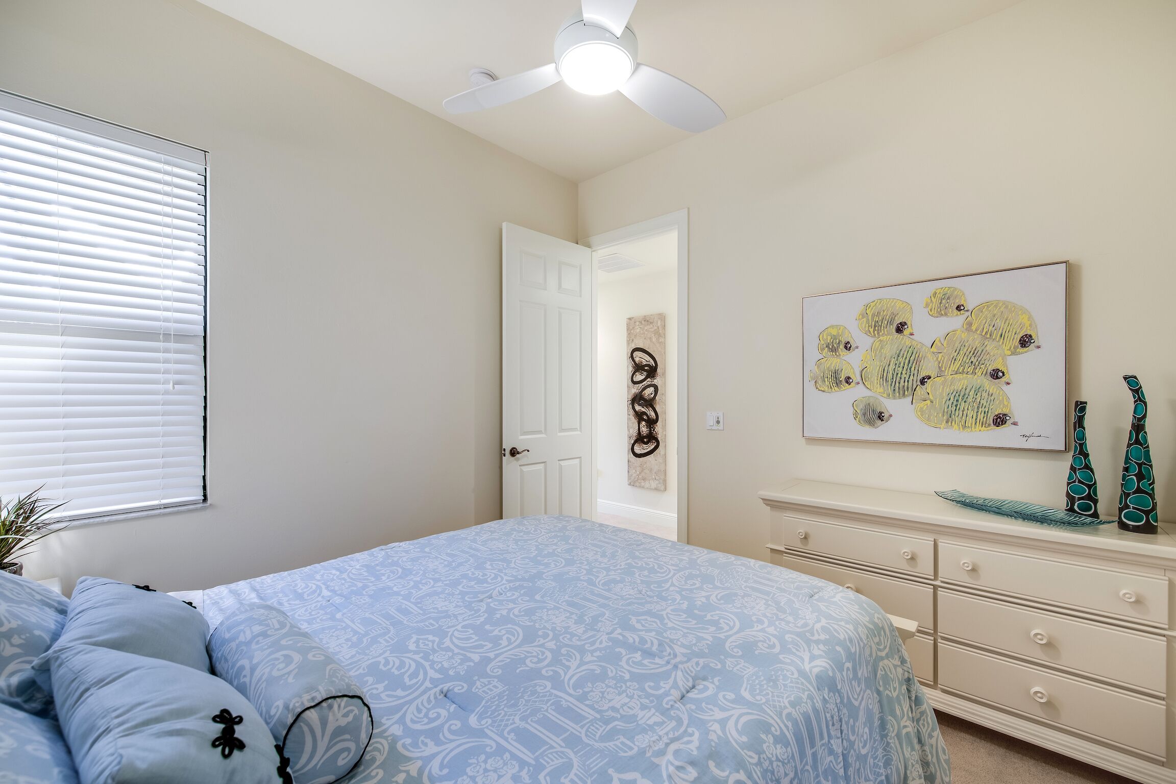 A bedroom with a bed, a ceiling fan, a door leading to another room, and a wall with a framed artwork.