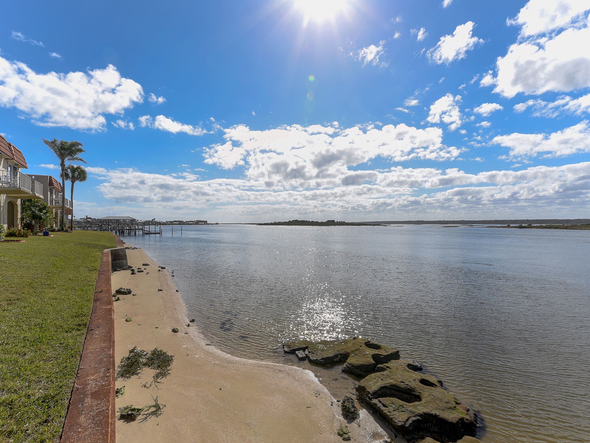 A waterfront property with a sandy beach, a pier extending into the water, and a house with a balcony overlooking the scene.