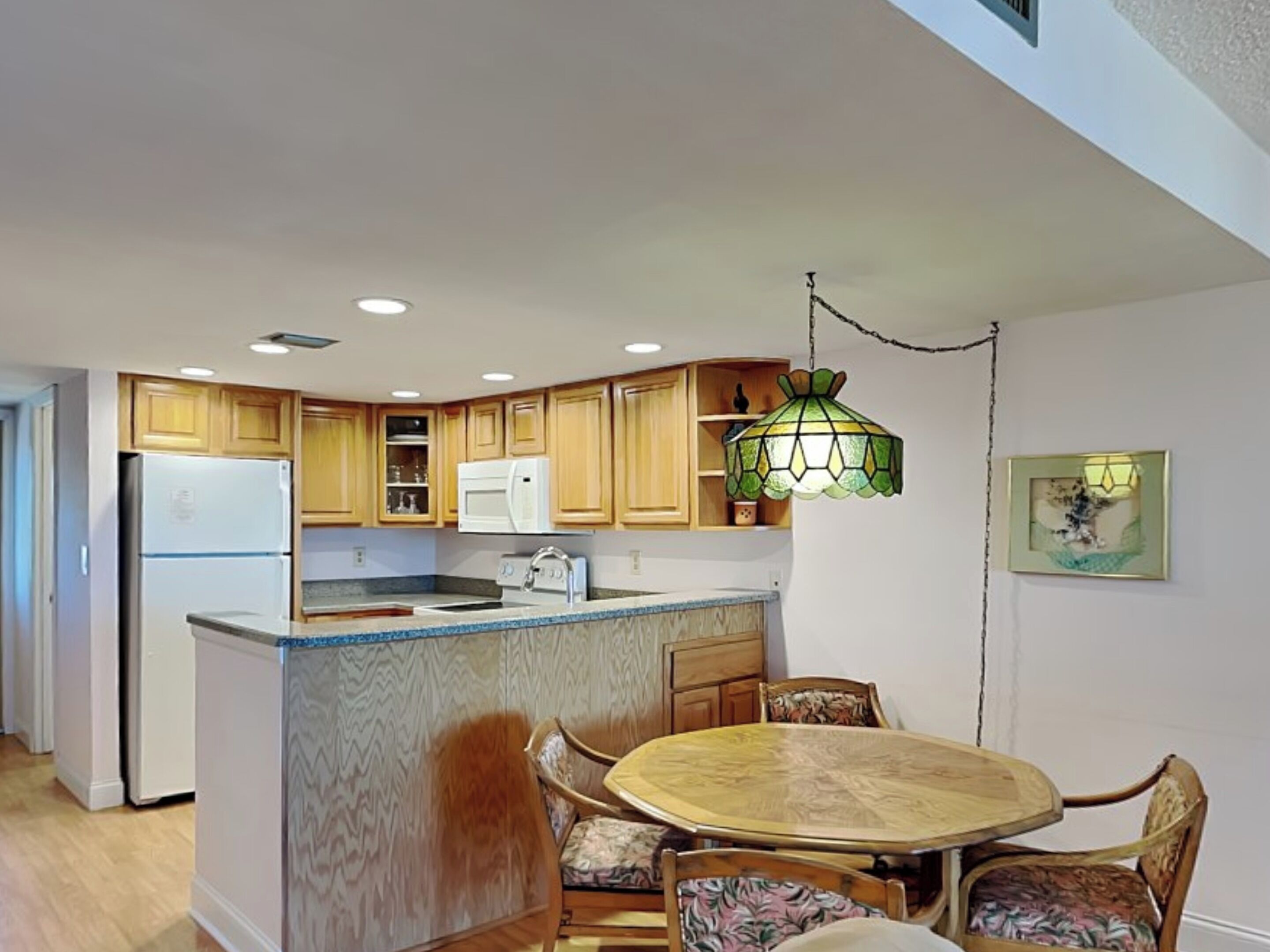 A compact kitchen with wooden cabinets, a central island with seating, and a hanging green pendant light.