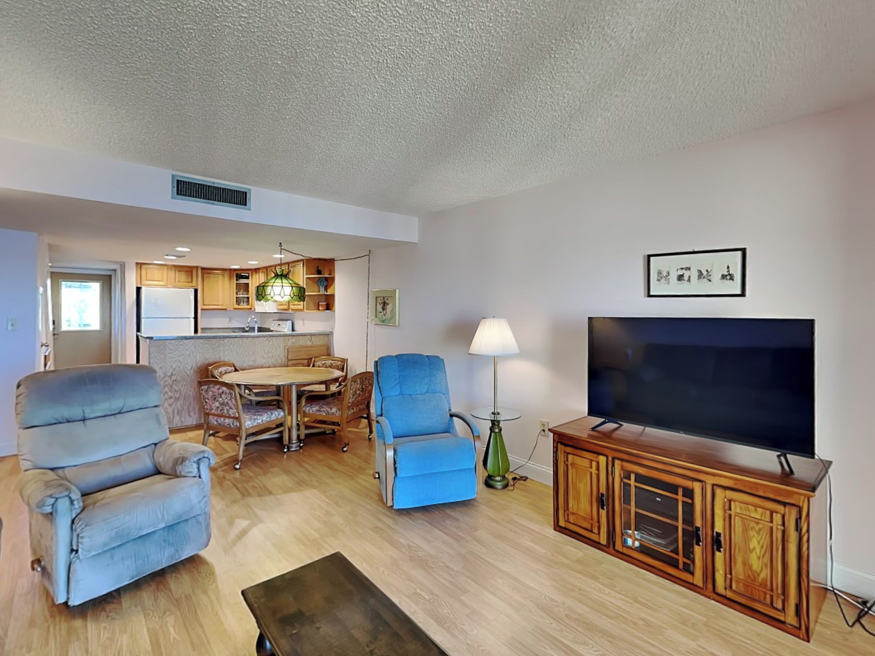 A living room with a TV, a blue sofa, a dining table with chairs, and a kitchen area in the background.