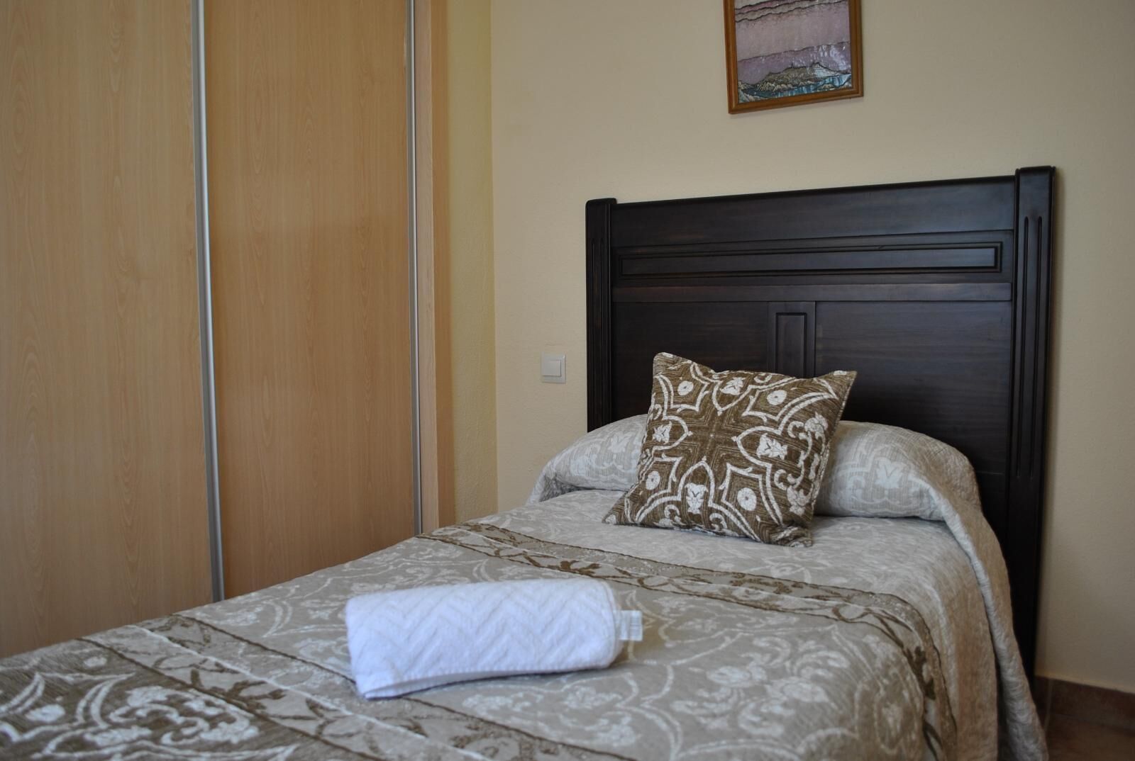 Una cama bien hecha con ropa de cama estampada y una almohada decorativa, un armario de madera y un cuadro en la pared.