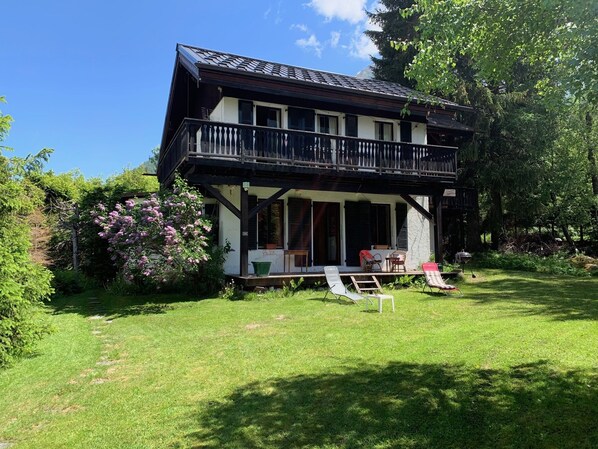 Une maison à deux étages dotée d’un balcon, entourée d’une végétation luxuriante et de plantes en fleurs.