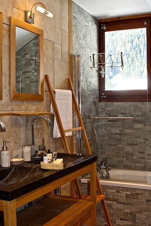 Une salle de bain avec un mur en pierre, un meuble-lavabo en bois, une baignoire et une fenêtre donnant sur la forêt.