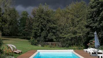 A rectangular swimming pool with a wooden deck surrounding it, set against a backdrop of dense trees and a cloudy sky.