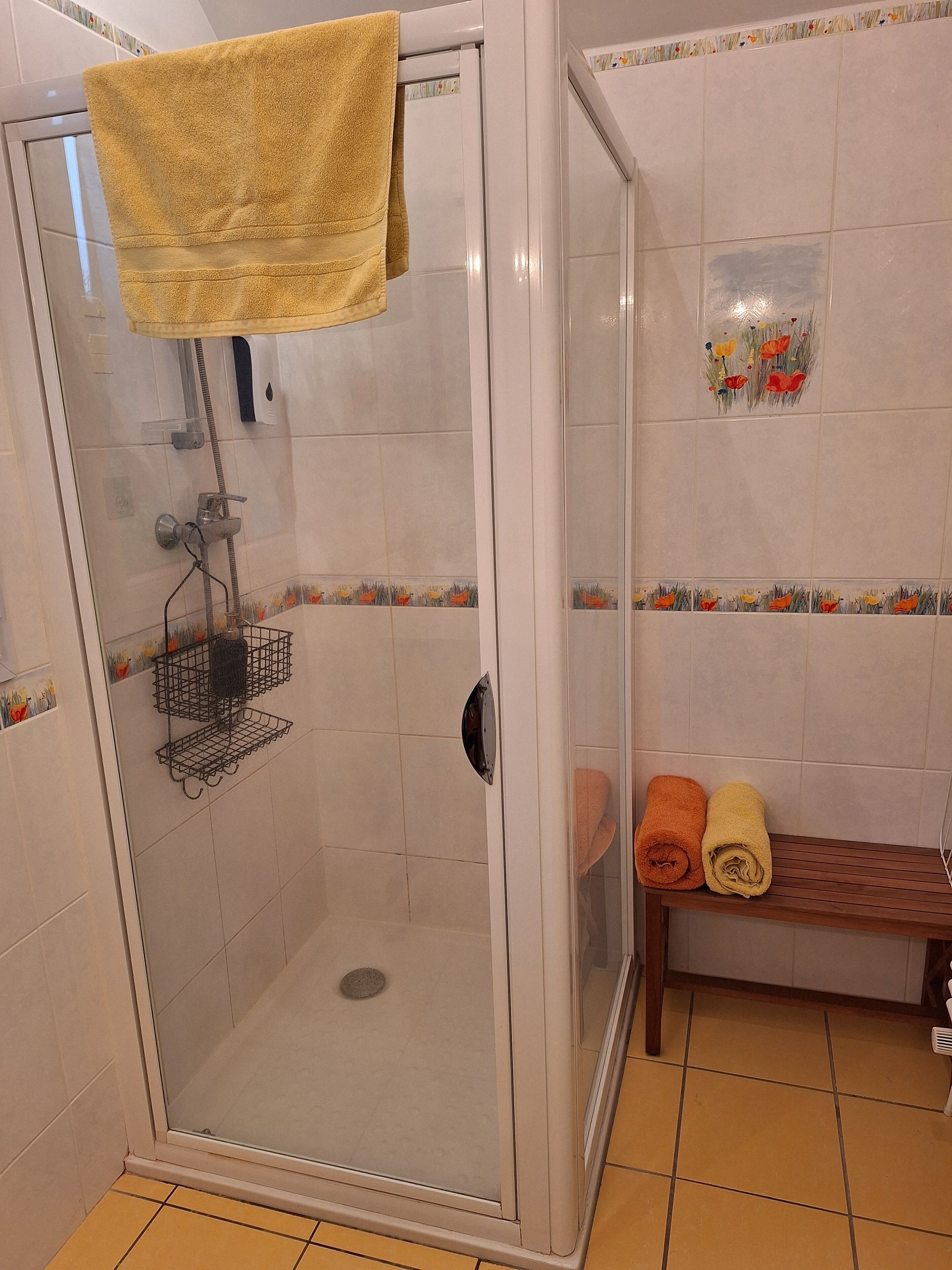 Una zona de ducha con puerta de vidrio, un perchero con una toalla amarilla y un banco con dos toallas enrolladas.