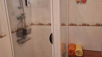 Una zona de ducha con puerta de vidrio, un perchero con una toalla amarilla y un banco con dos toallas enrolladas.