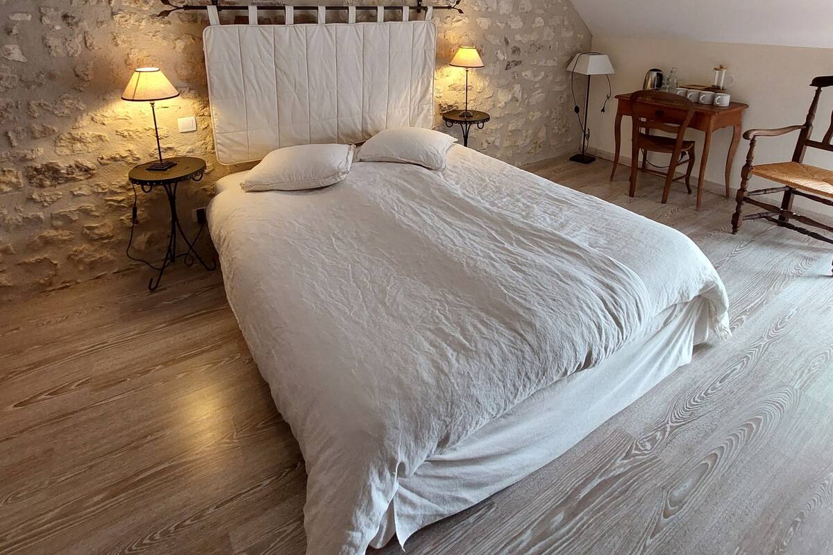 Une chambre avec un plafond en pente, des murs en briques apparentes, un plancher en bois, un lit avec du linge de lit blanc, deux lampes de chevet et une petite table en bois avec des chaises.