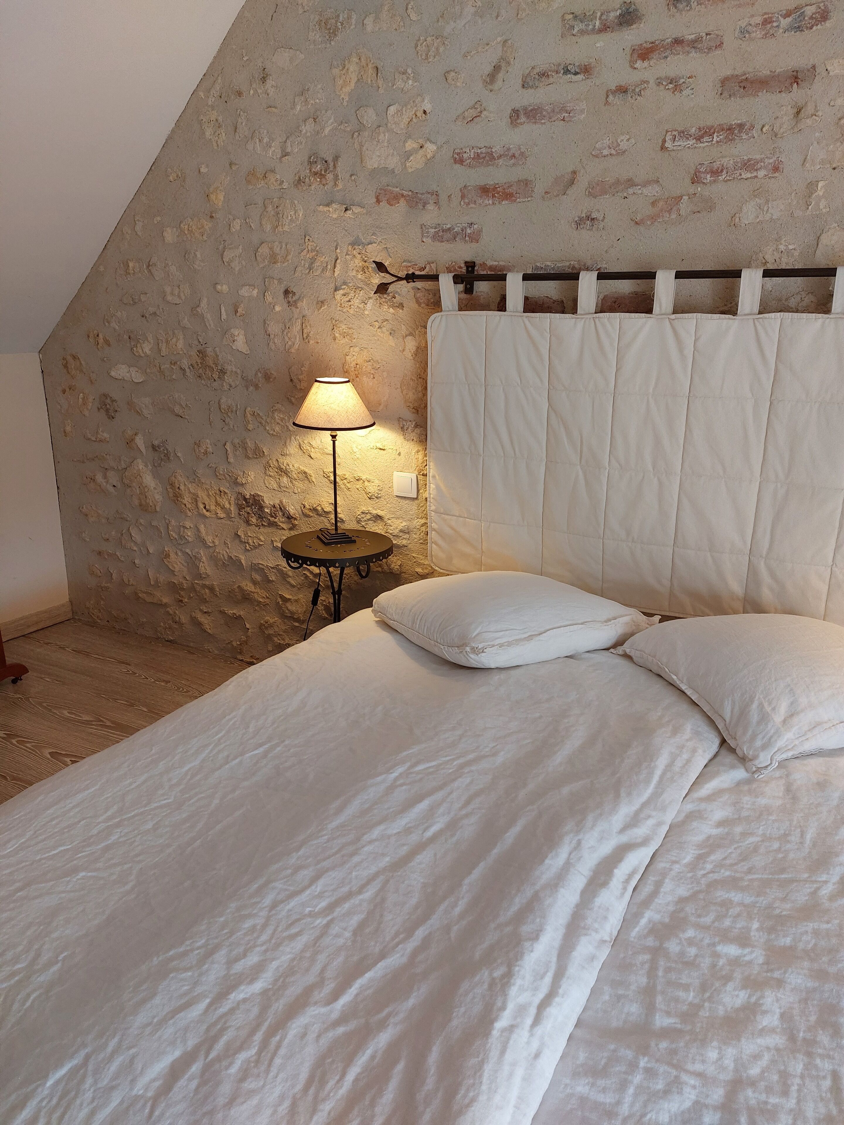 A bedroom with a sloped ceiling, a bed with white bedding, a bedside lamp, and a brick wall.