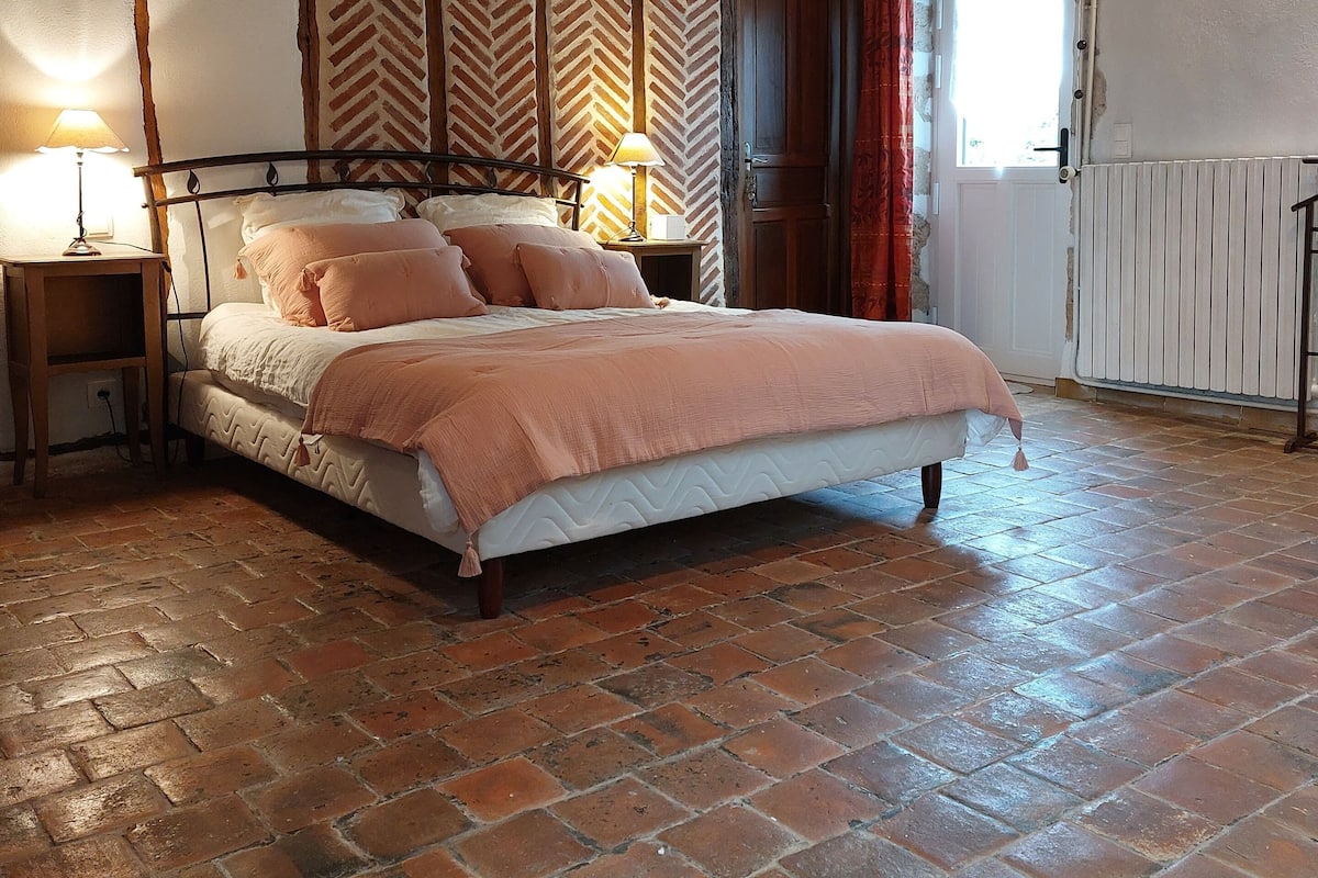 Une chambre à coucher avec un lit, des poutres en bois, un mur orné d’un motif en chevrons, un radiateur et une fenêtre avec des rideaux.