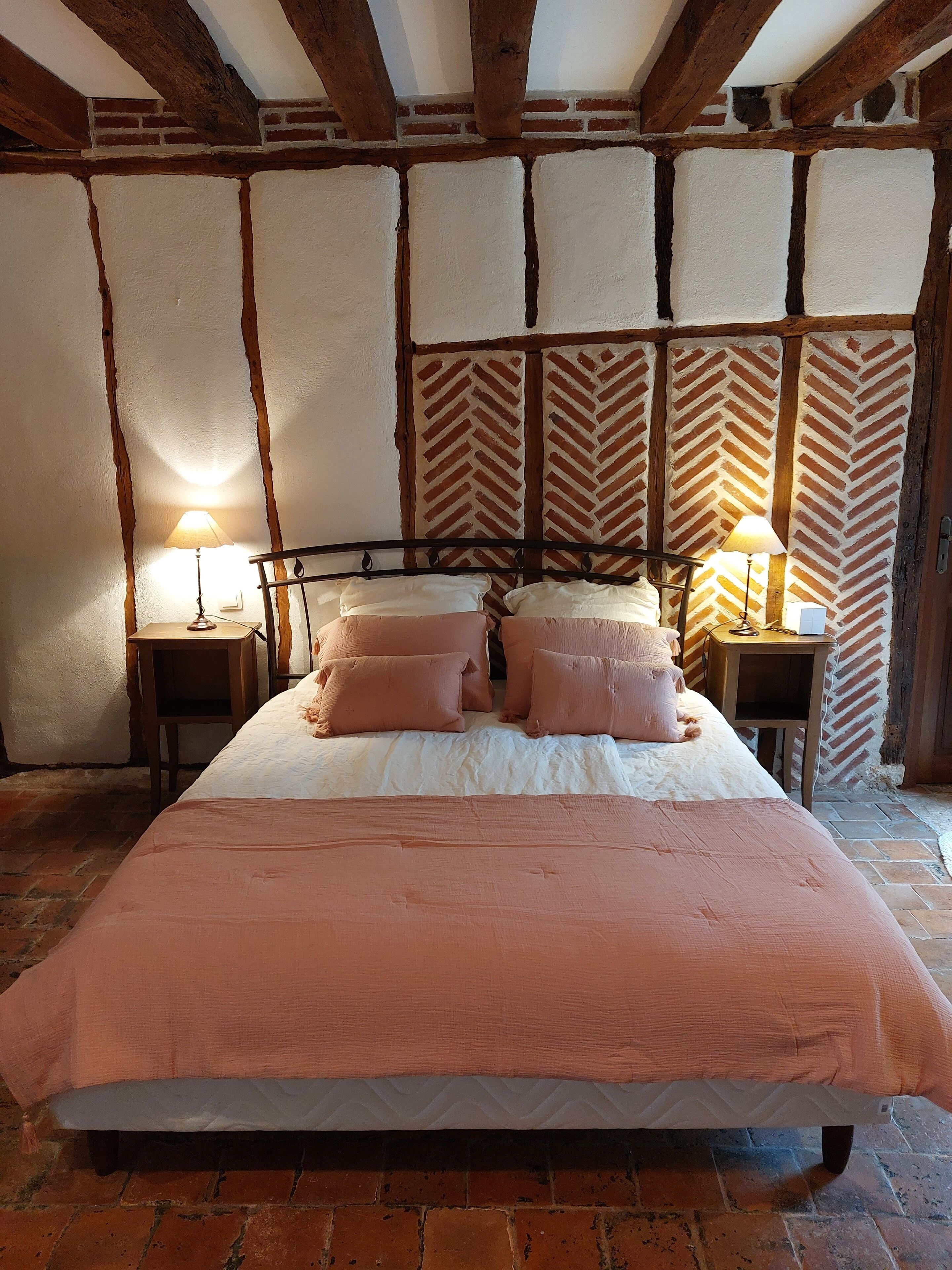 Een bed met een roze dekbed en kussens, houten balken, een bakstenen muur en twee bedlampen.