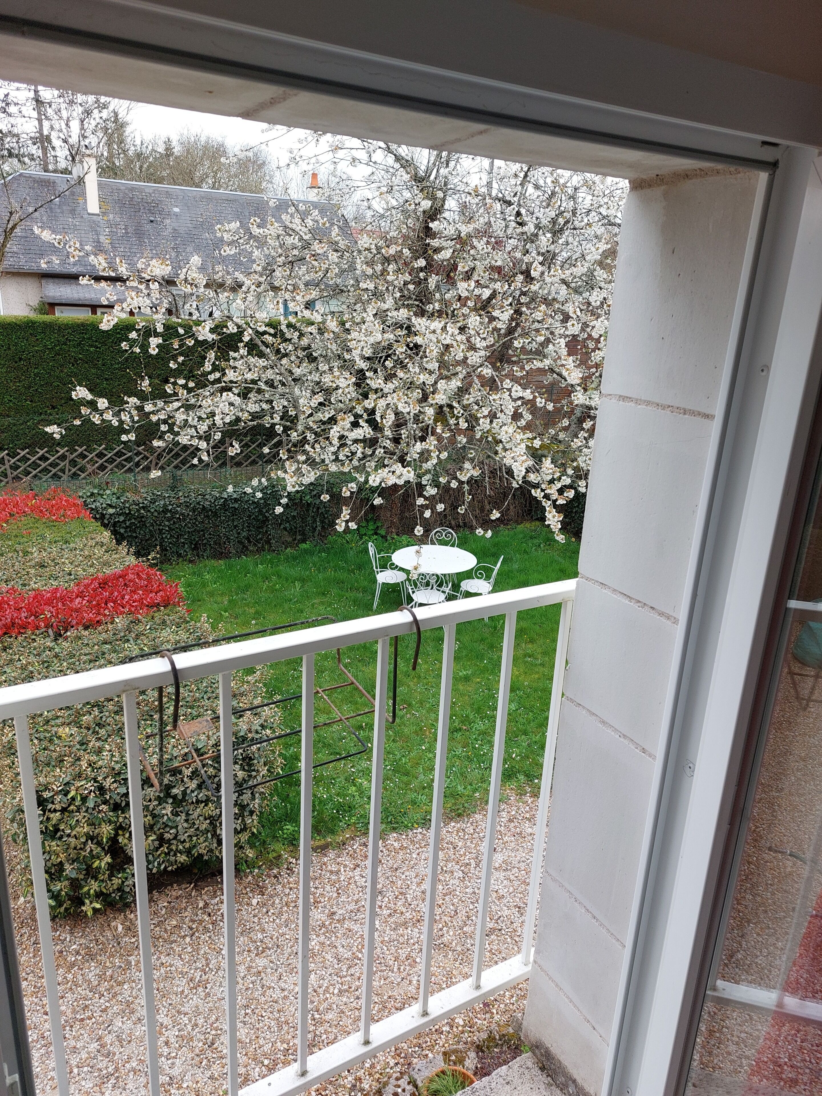 A balcony with a view of a garden, a white table, and a hedge.