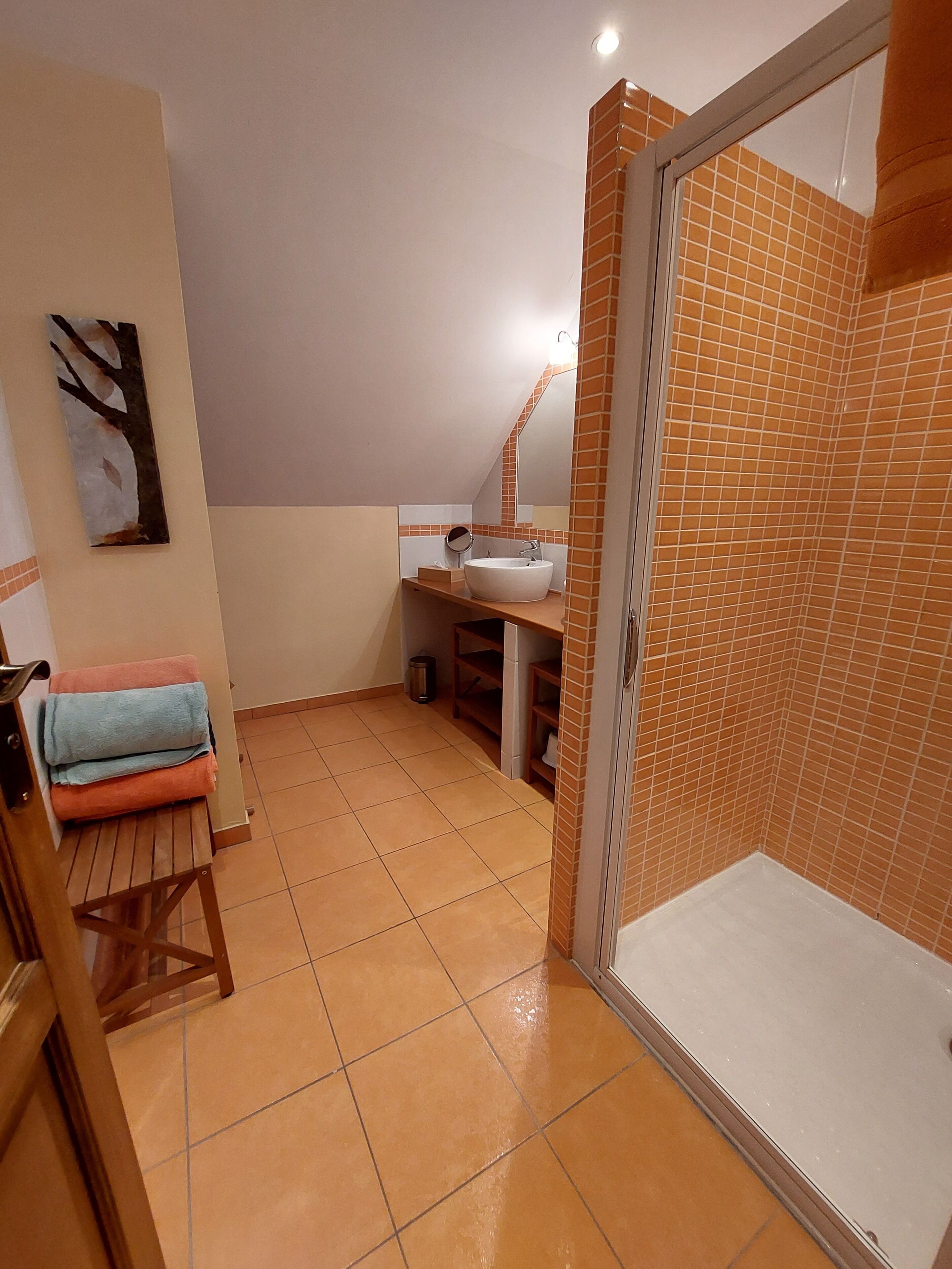 A bathroom with a white sink, a mirror, and a shower area with orange tiles. There is a towel rack with folded towels and a trash can.