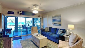 A spacious living room with a blue sofa, a patterned armchair, and a coffee table. There is a large window offering a view of the outdoors.