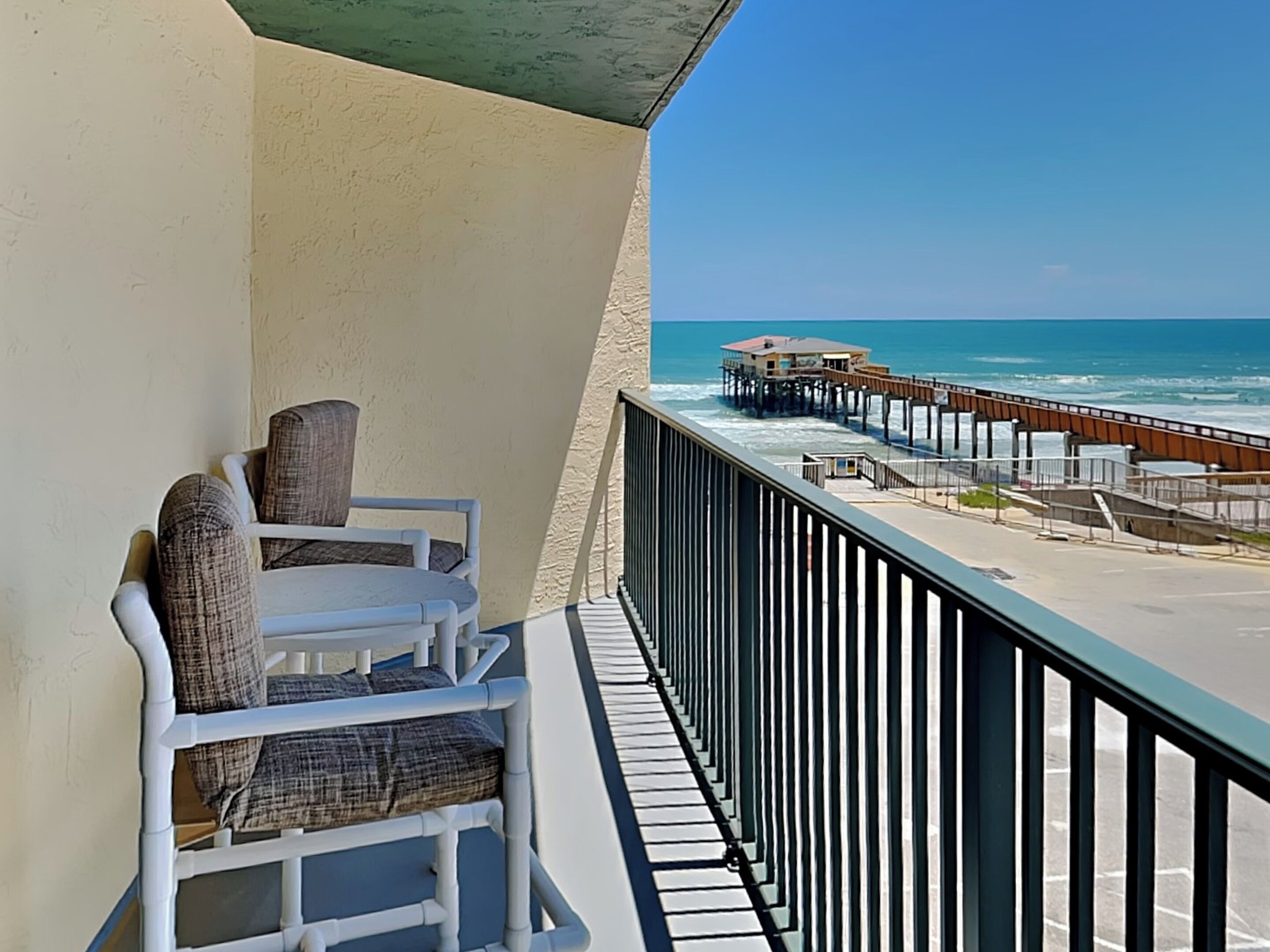 A balcony with white chairs and a table, overlooking a beach with a pier and ocean.