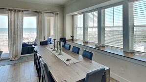 A spacious dining area with a long wooden table, blue chairs, and a view of the beach through large windows.
