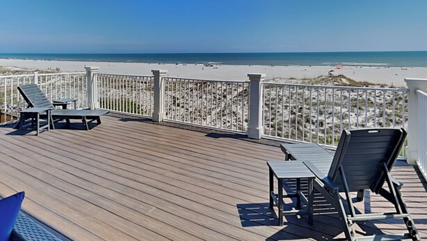 A wooden deck with two lounge chairs and a table, overlooking a beach with white sand and a clear blue sky.