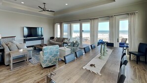 A spacious living room with a large wooden dining table, comfortable seating, a flat-screen TV, and a view of the ocean through large windows.