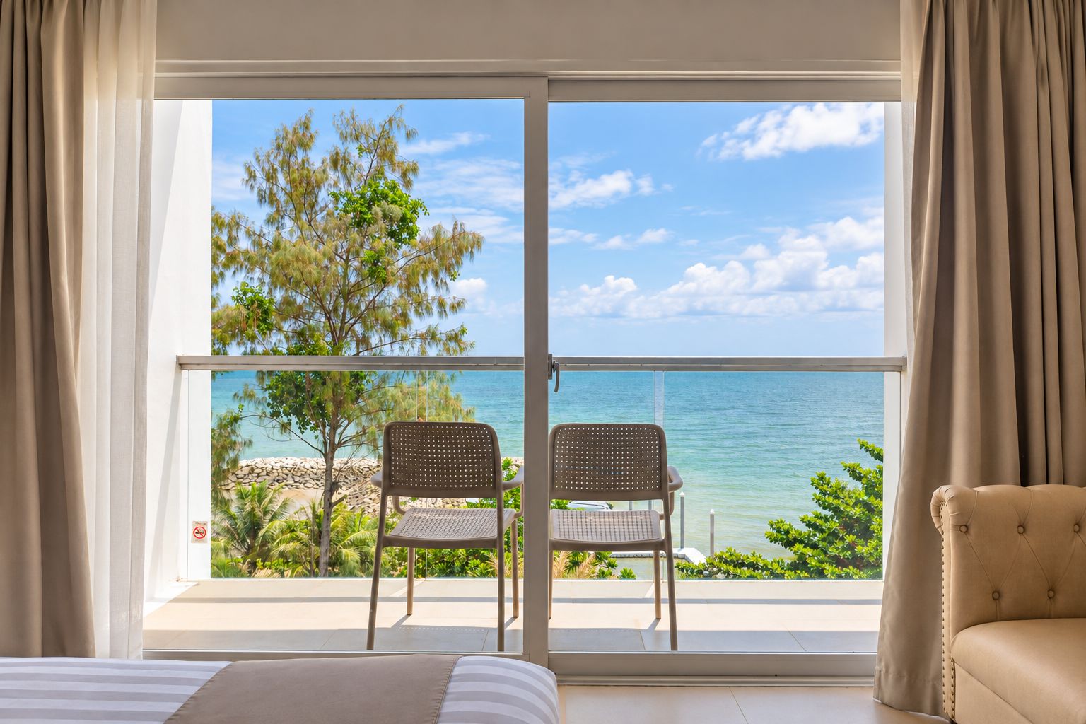 A room with a bed, two chairs, and a view of the sea through a large glass door.
