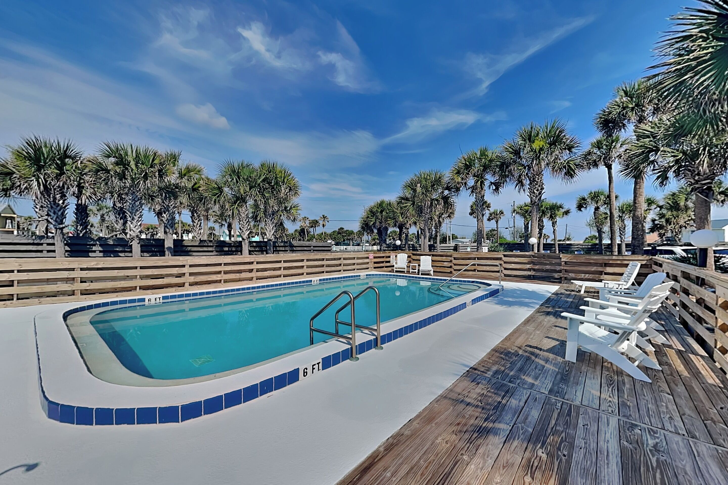 A swimming pool with a wooden deck, surrounded by palm trees and a clear sky.