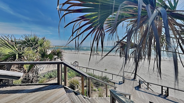 Une terrasse en bois avec une rambarde, donnant sur une plage bordée de palmiers et sur l’océan.