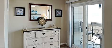 A room with a white dresser, a mirror, and two framed pictures on the wall. There is a sliding glass door leading to a balcony with outdoor furniture.
