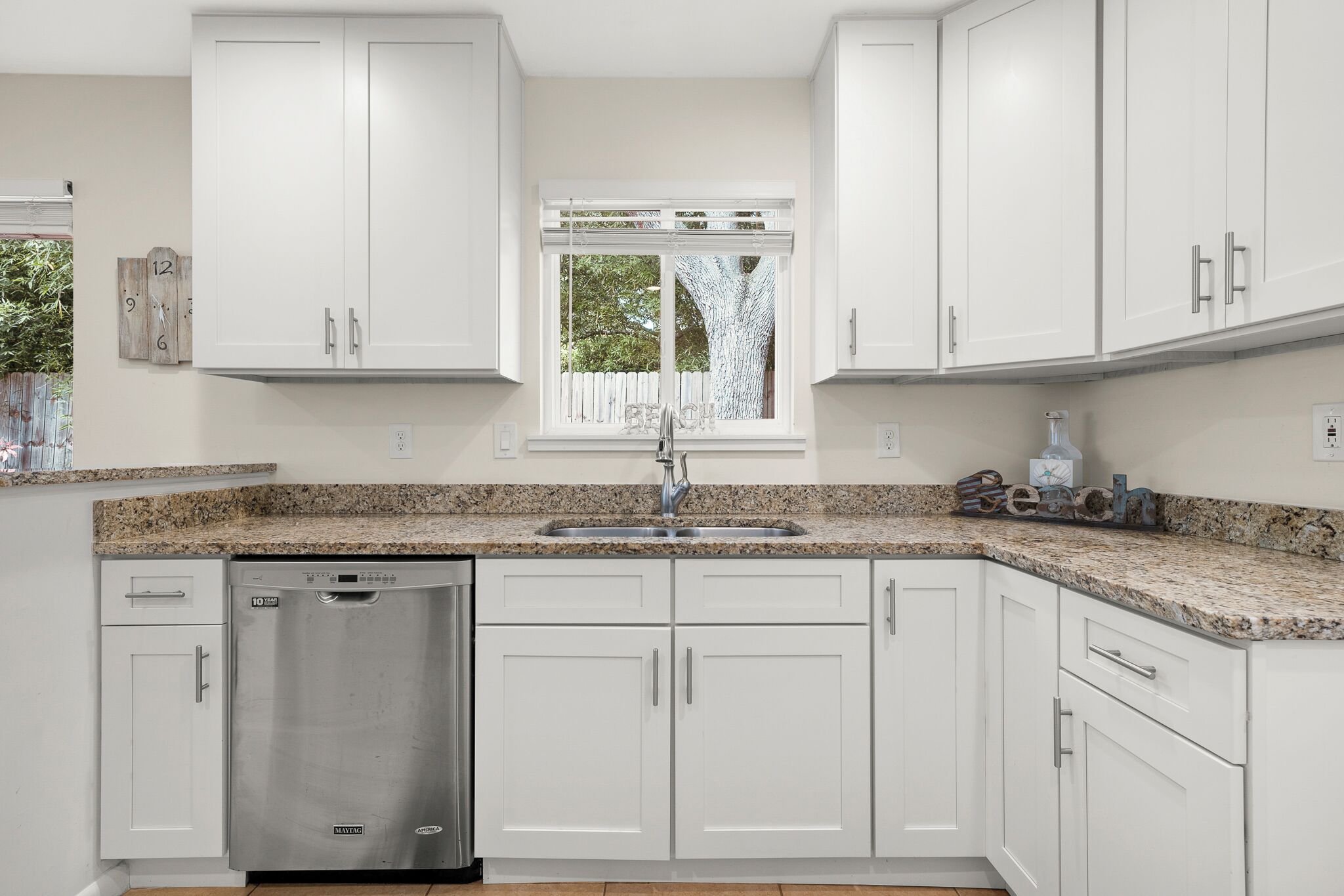 A modern kitchen with white cabinets, a granite countertop, a stainless steel dishwasher, and a window with a view of trees.