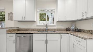 A modern kitchen with white cabinets, a granite countertop, a stainless steel dishwasher, and a window with a view of trees.