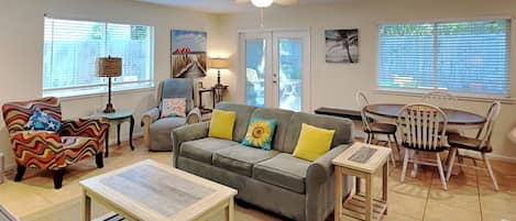 Una sala de estar con un sofá gris, un sillón colorido, una mesa de centro de madera y un comedor con mesa y sillas de madera.
